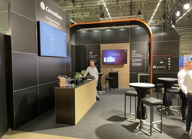 A modern exhibition booth with black and wood accents displays the Consilium logo. Two people stand near high tables and screens, with brochures and plants on the counter, under bright overhead lights.