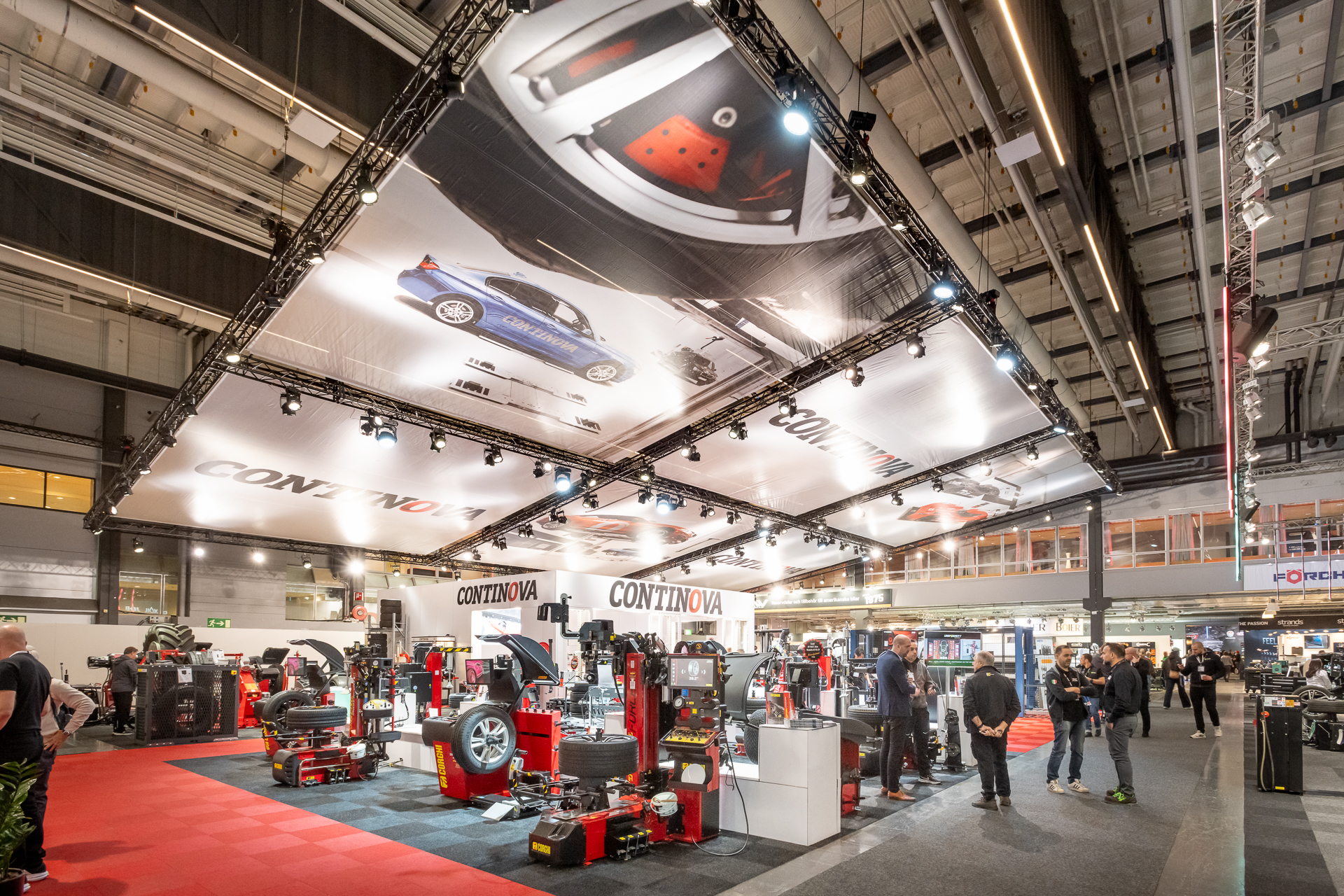 A large, well-lit exhibition hall with automotive equipment displays, large overhead banners featuring car images, and people walking and interacting with the exhibits. The booth is marked Continova.