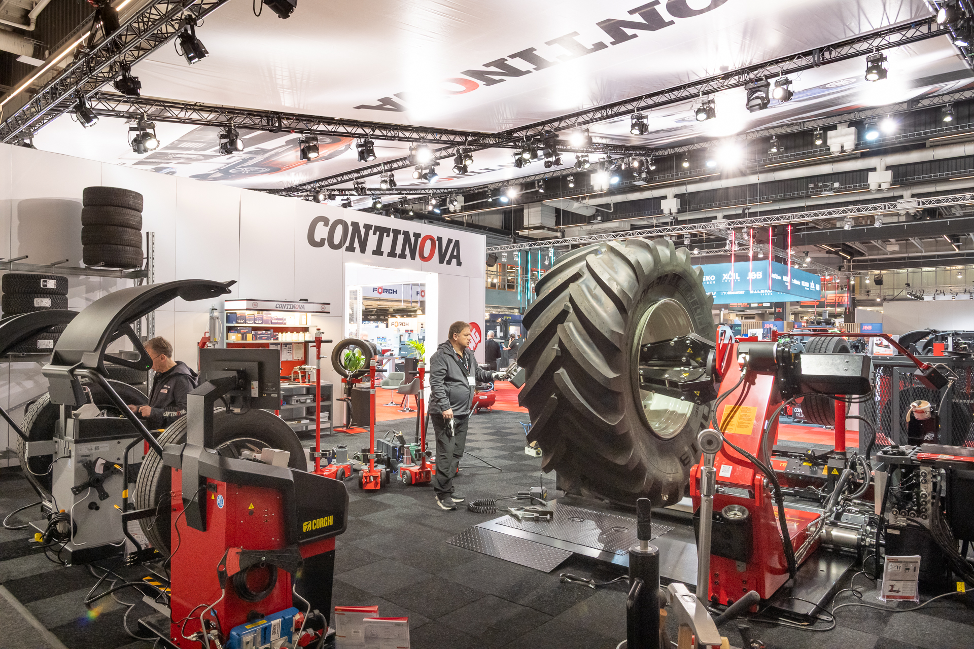 A trade show booth for CONTINOVA features large heavy-duty tire equipment, a giant tractor tire on a machine, various tools, display shelves, and two people working under bright lights.