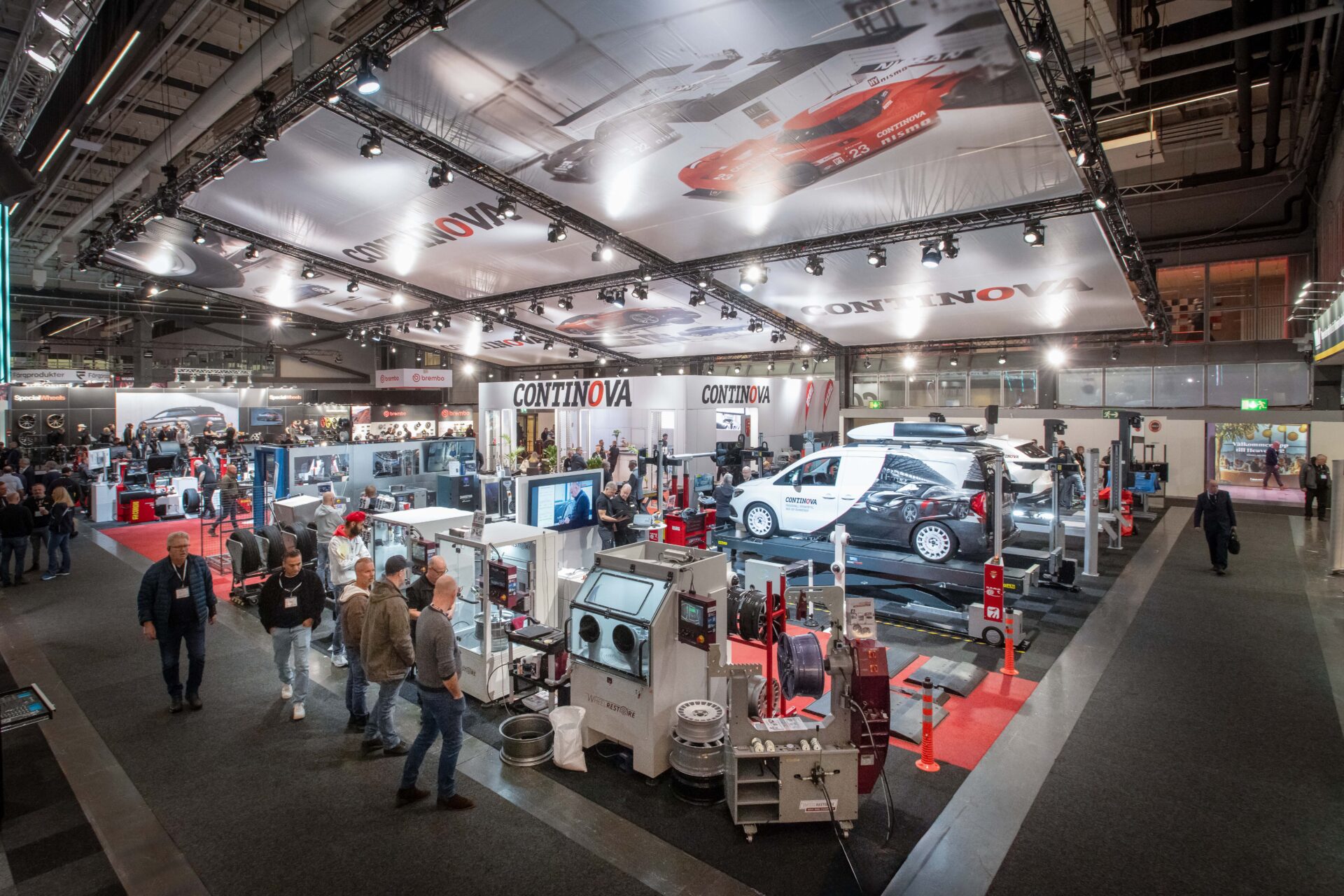 A busy trade show or expo hall features various automotive displays, machinery, and equipment booths, with people walking around and examining exhibits under large banners reading CONTINOVA.