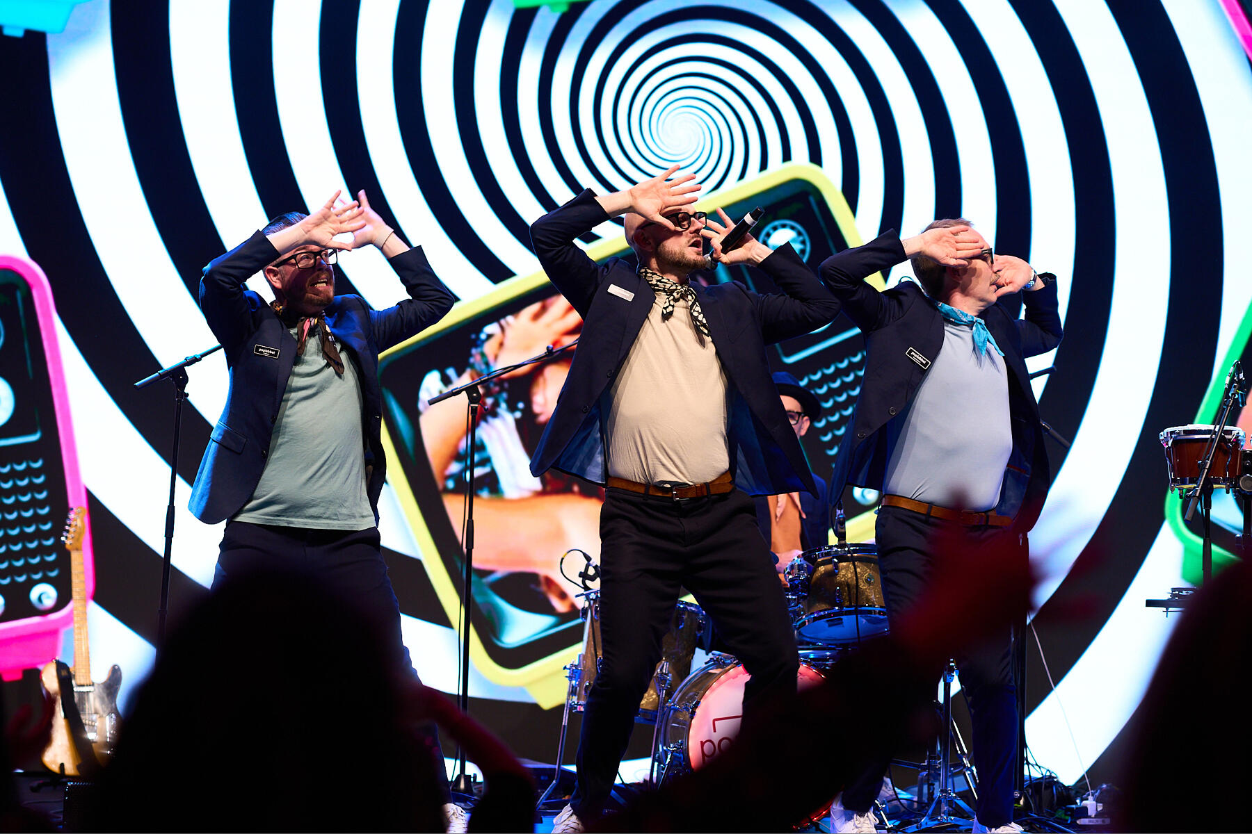 Three men in suits perform energetically on stage with their hands on their foreheads. A colorful, hypnotic spiral and retro graphics fill the background, and musical instruments are visible behind them.