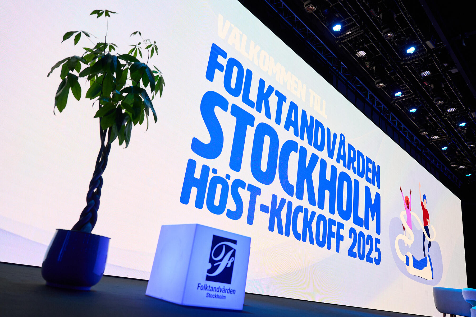 A stage with a large screen displays “Välkommen till Folktandvården Stockholm Höst-Kickoff 2025” with blue text and cartoon figures; a potted plant and a bench with the Folktandvården Stockholm logo are in front.