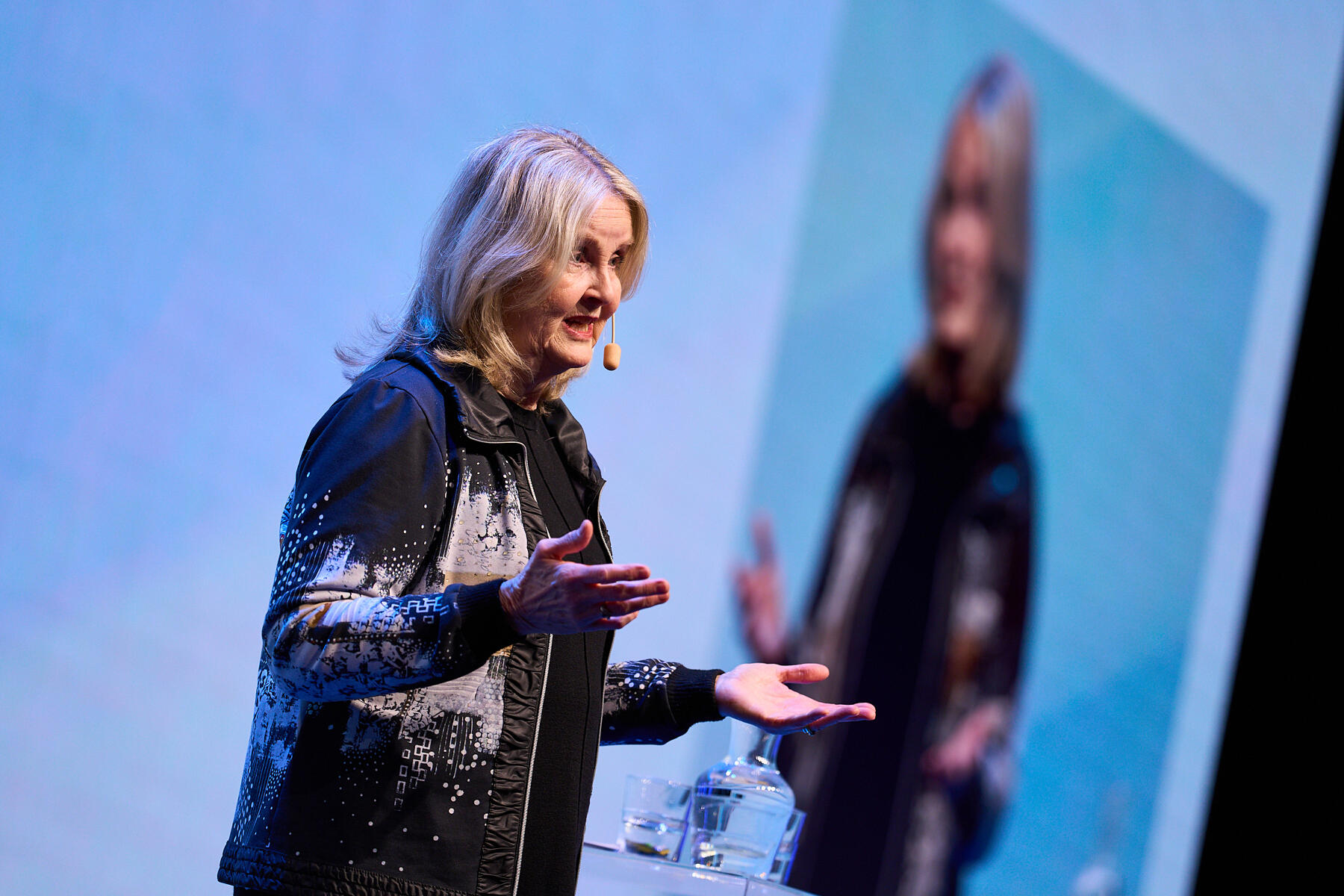 A woman with long blonde hair speaks on stage with a headset microphone, gesturing with her hands. She wears a black and white patterned jacket and stands in front of a large projected image of herself.