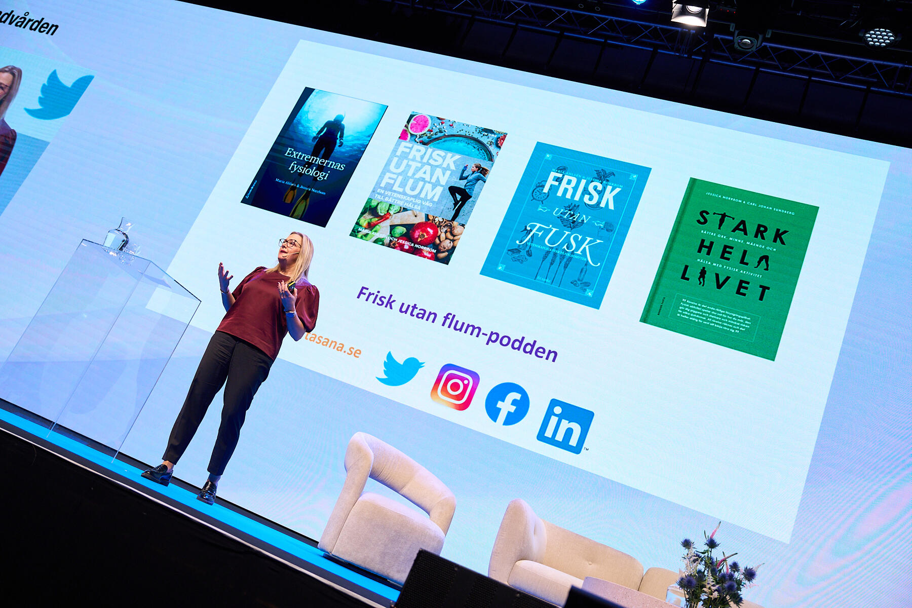 A woman stands on stage giving a presentation. Behind her is a large screen displaying four book covers and social media icons for Instagram, Twitter, Facebook, and LinkedIn.