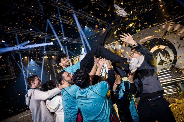 A group of people in teal jerseys lift a teammate into the air while gold confetti falls around them on a brightly lit stage, celebrating their victory.