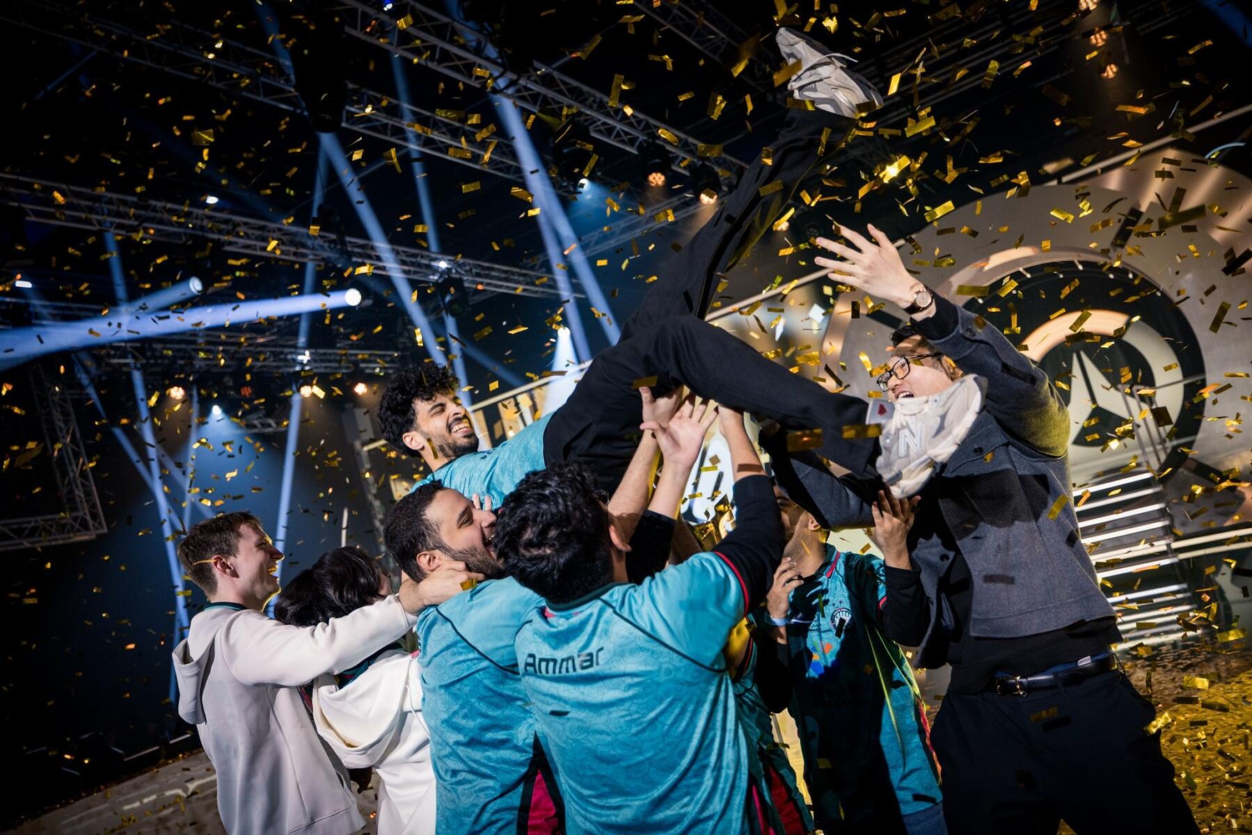 A group of people in teal jerseys lift a teammate into the air while gold confetti falls around them on a brightly lit stage, celebrating their victory.