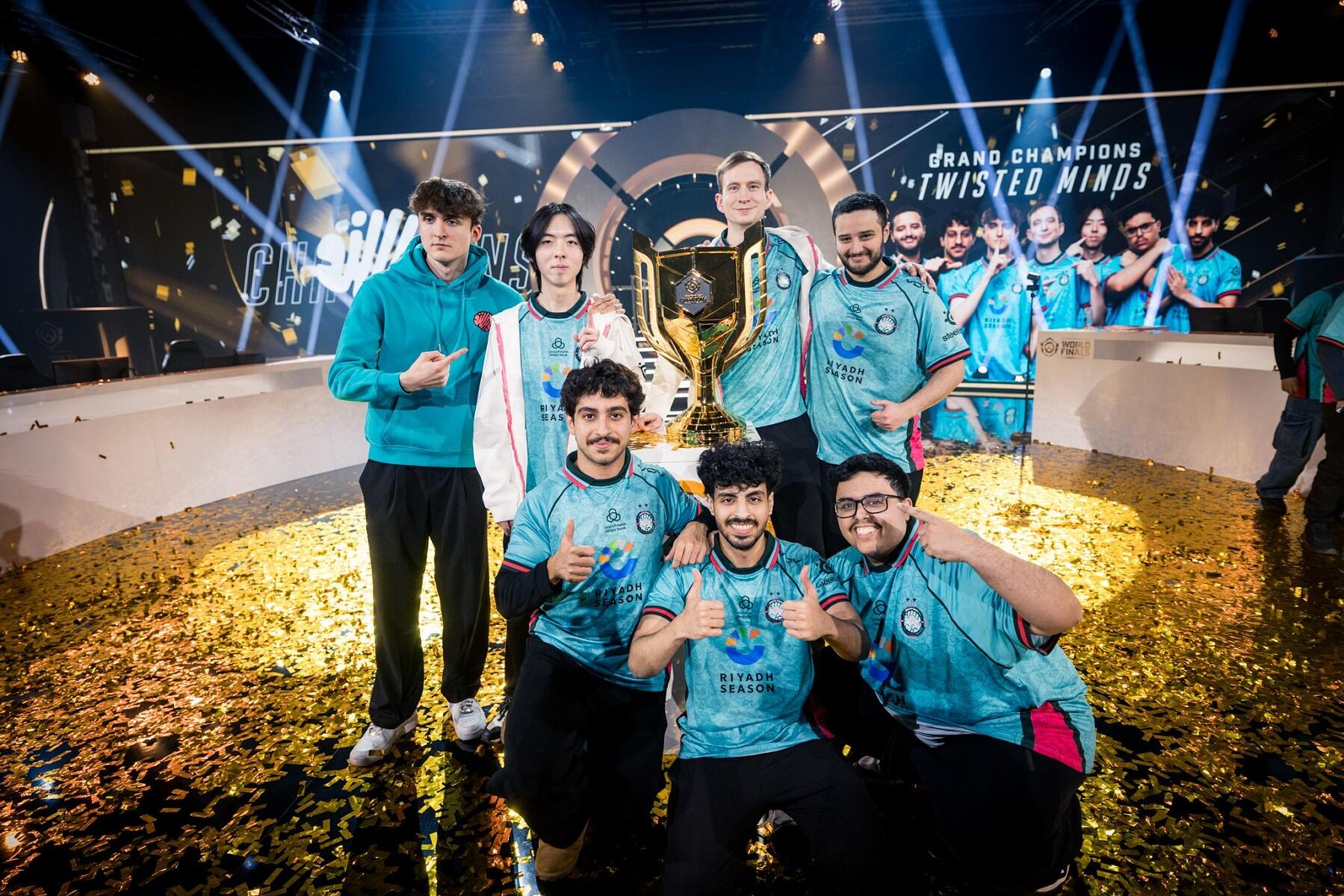 A group of seven esports players in teal jerseys pose and smile on a confetti-covered stage, celebrating their championship win with a trophy, bright lights, and a large “Twisted Minds” sign in the background.