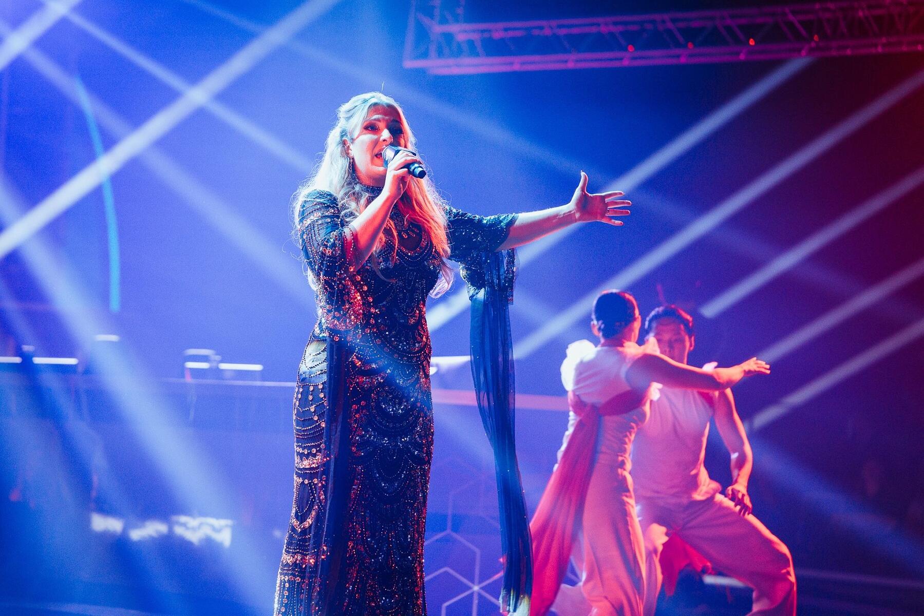 A woman in a sparkling dress sings passionately on stage, holding a microphone, with colorful spotlights and two backup dancers performing behind her. The atmosphere is vibrant and energetic.