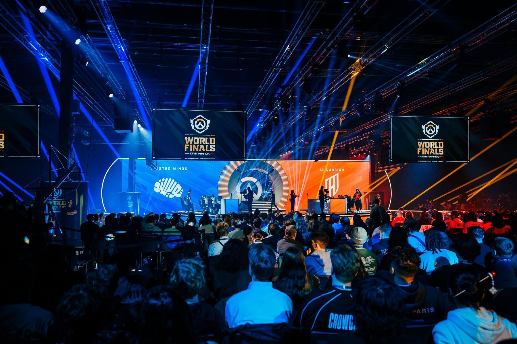 A large audience watches an esports world finals event in a brightly lit arena, with players seated on stage and large screens displaying game graphics and team logos above them.