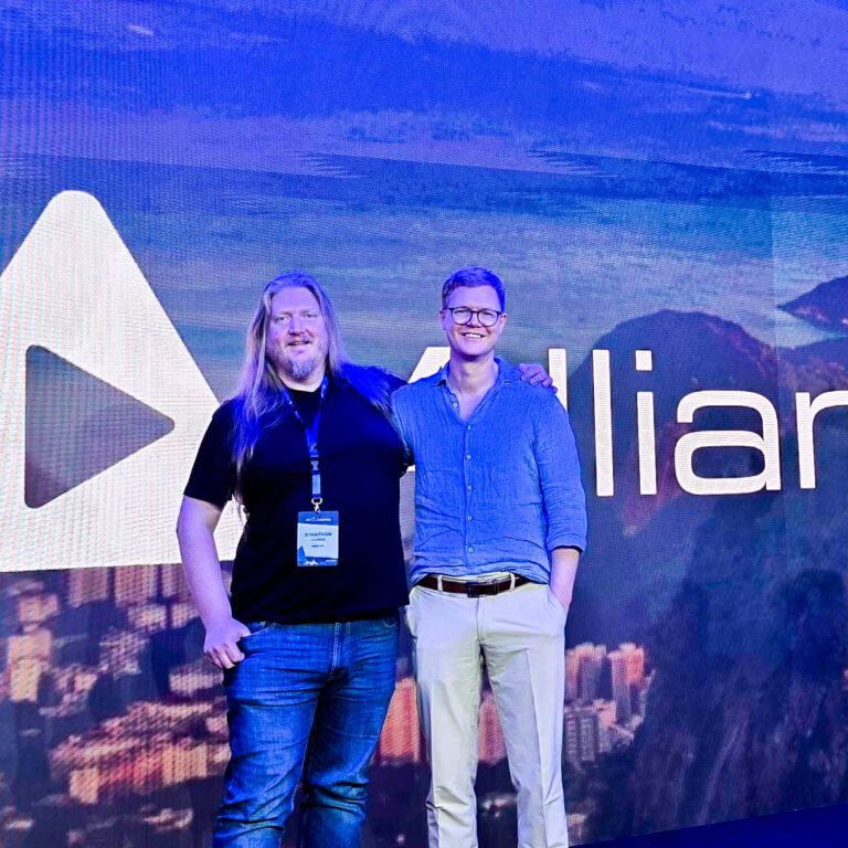 Two men stand smiling in front of a large screen displaying the word Allian and a mountain landscape. One man has long blond hair and wears a black shirt and jeans; the other wears glasses, a blue shirt, and light pants.