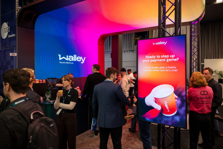 A group of people gathers at a vibrant expo booth with colorful lights and a large digital sign that reads, Ready to step up your payment game? alongside an image of a coffee cup. The booth is branded with Walley.