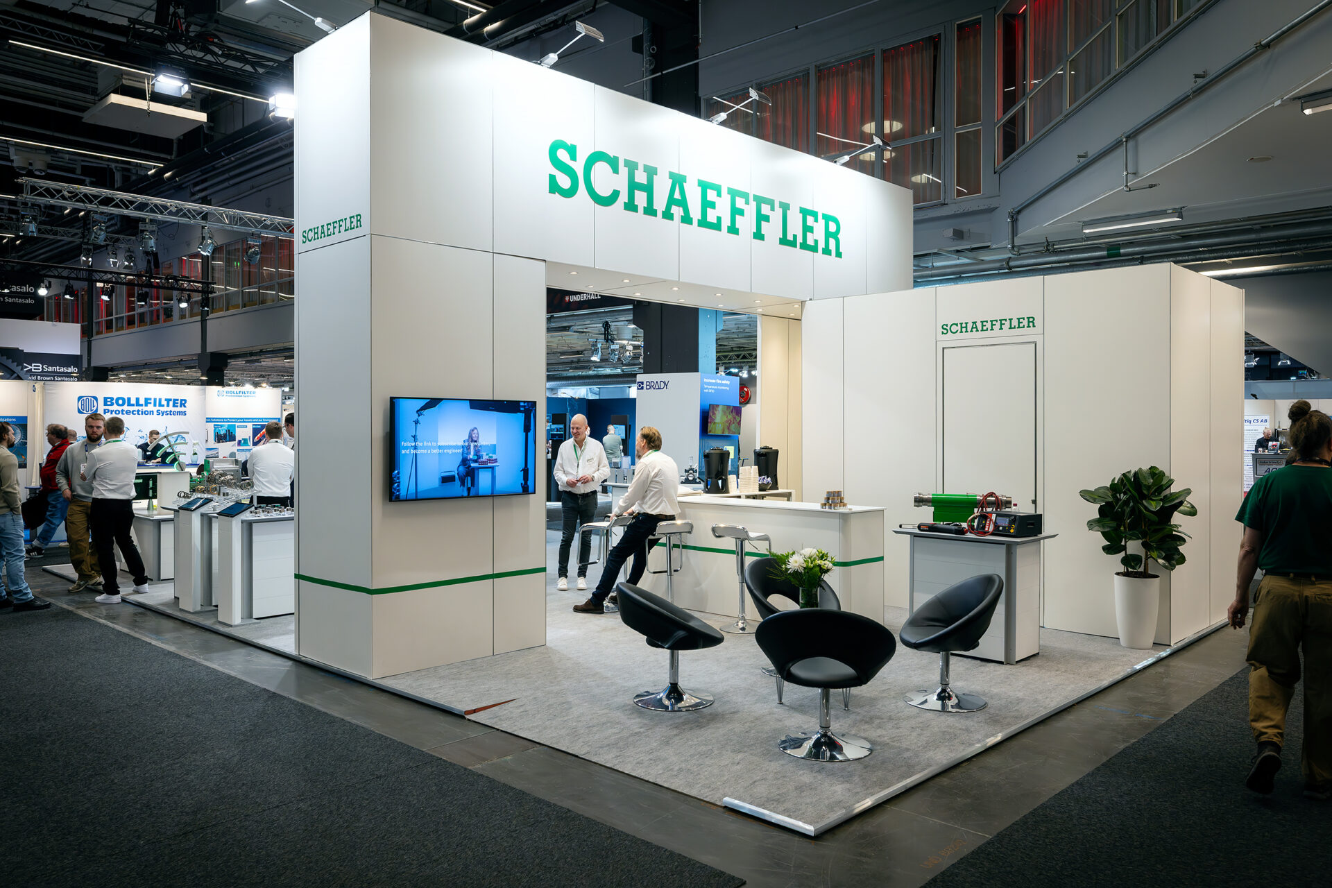 A modern exhibition booth for Schaeffler with a white and green color scheme, chairs, display screens, plants, and people interacting, set in a large, well-lit convention hall.