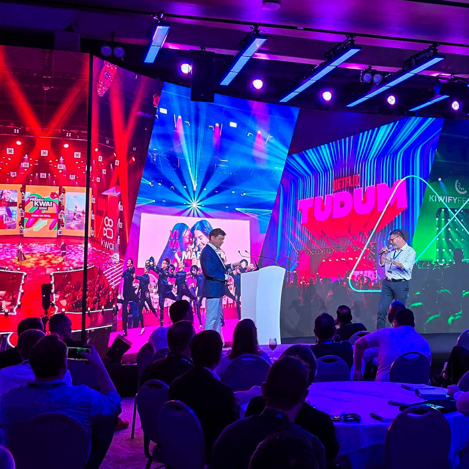 A brightly lit conference with two speakers on stage, colorful screens displaying logos and graphics, and an audience seated at round tables, watching and taking photos.
