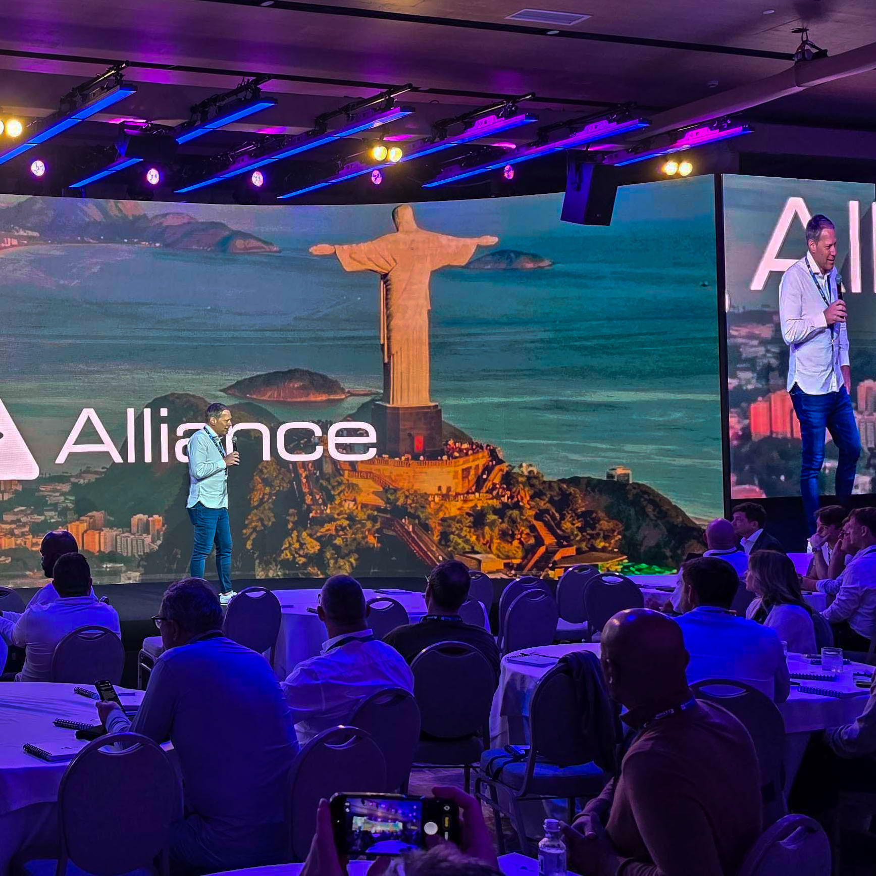 A man stands on stage giving a presentation to an audience. Behind him is a large screen displaying the Christ the Redeemer statue in Rio de Janeiro, with the word “Alliance” superimposed on the image.