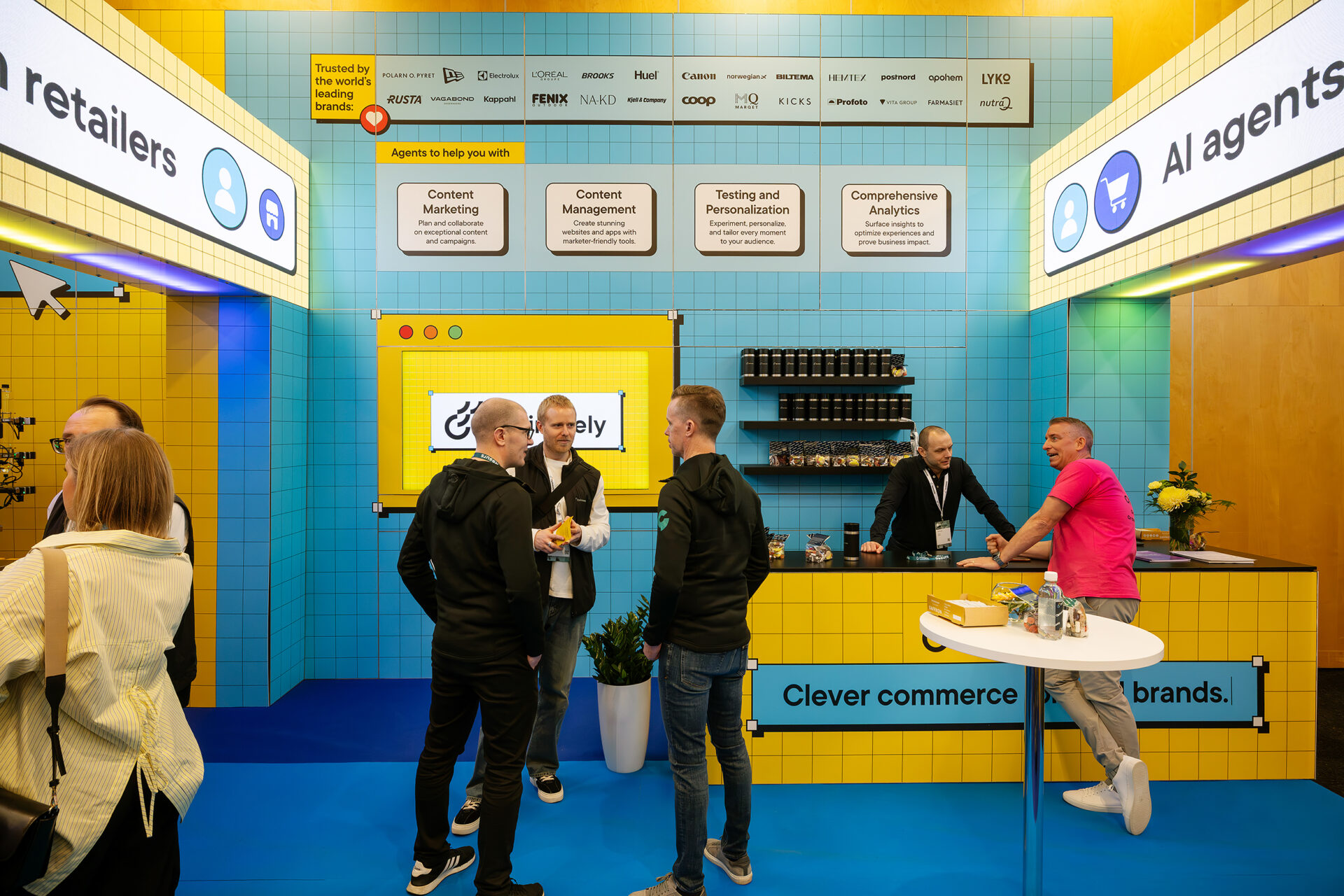 People interact at a brightly colored trade show booth with blue and yellow tiled walls, featuring signs about AI agents and retailers. The booth has a counter, displays, and branding materials.