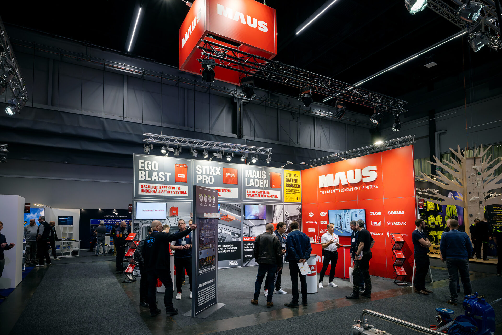 A trade show booth displays MAUS-branded banners and fire safety products. Several people are interacting with staff and looking at informational displays under bright overhead lighting.
