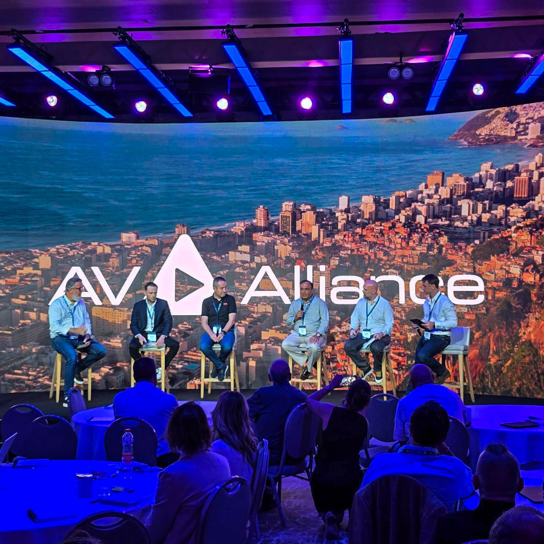 Six people sit on a stage panel under purple lights, with a large screen behind them displaying AV Alliance and an aerial cityscape. Audience members are seated at round tables in the foreground.