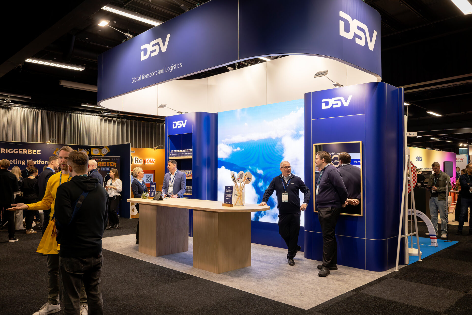 A trade show booth for DSV, a logistics company, features a large blue display with the DSV logo and a screen showing clouds. Several people are interacting around the booth in a busy exhibition hall.