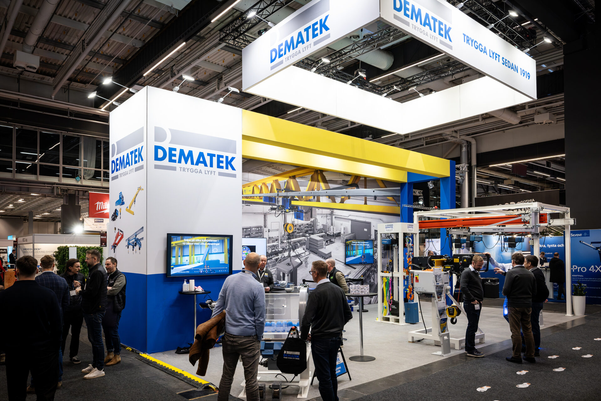 Trade show booth for DEMATEK with bright blue and yellow signage, screens displaying information, industrial equipment, and several people interacting and walking around the exhibit.