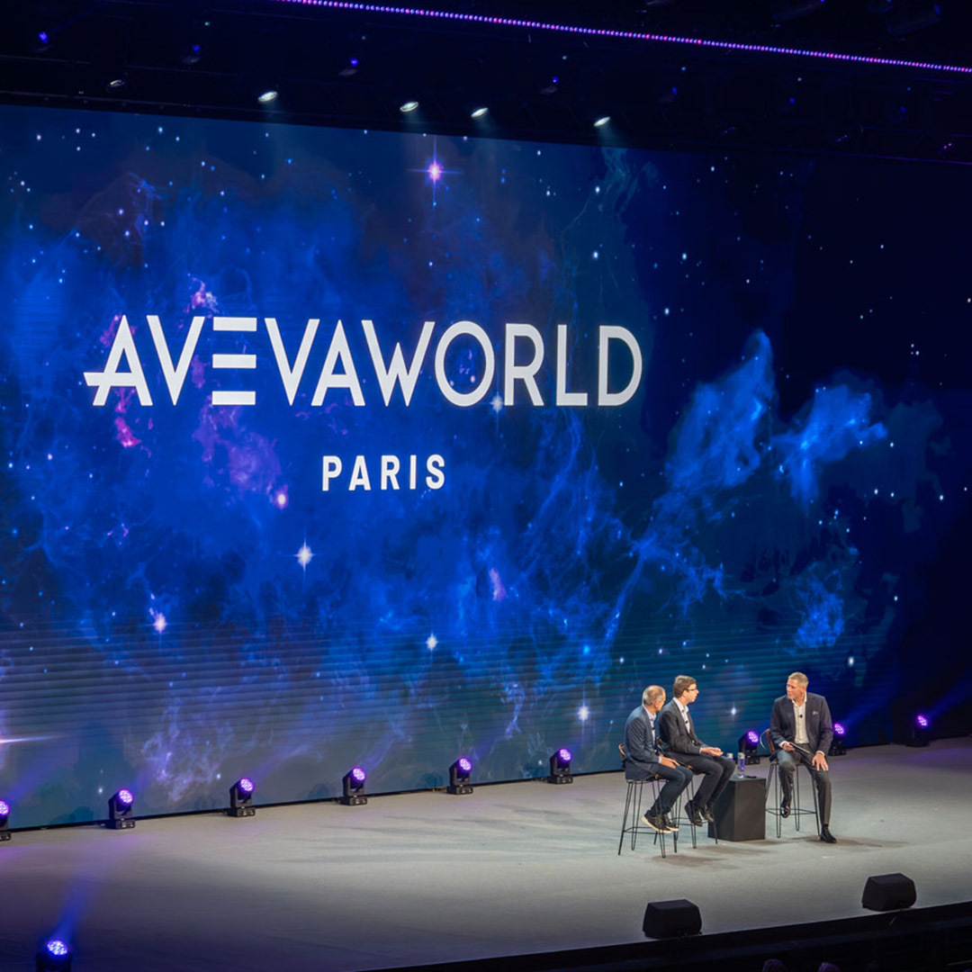 Three people sit on stage in front of a large screen displaying AVEVA WORLD PARIS with a blue, space-themed background. Stage lights illuminate the area as they engage in discussion.