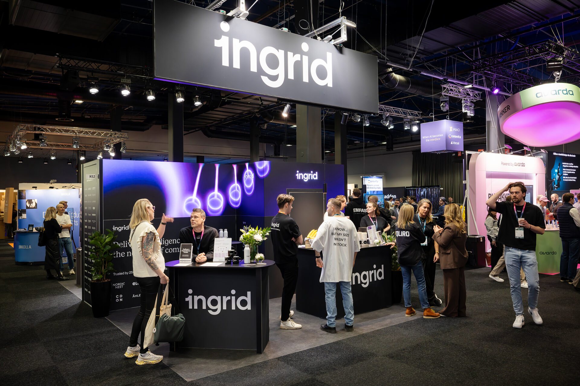 A busy exhibition booth for ingrid at a trade show, with staff engaging visitors, promotional materials on counters, and illuminated signs overhead. Other booths and attendees are visible in the background.