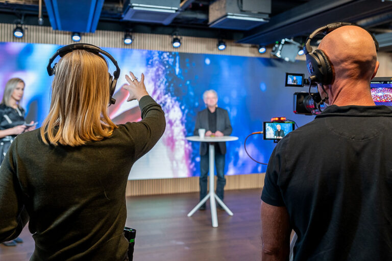 A woman with a headset gestures toward a man speaking at a small round table on a studio set, while a cameraman films the scene; colorful lights illuminate the background.