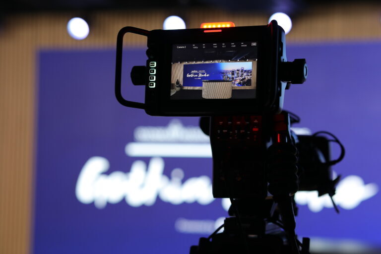 A video camera focuses on a blurred blue stage backdrop, with its screen displaying a clear image of the stage and the words Gastric Session. Bright lights are visible in the background.
