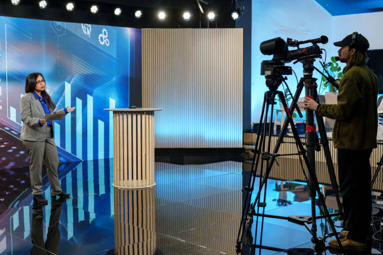 A woman in a suit stands on a studio set holding papers and speaking, while a cameraman films her. The background features blue graphics and vertical light panels.