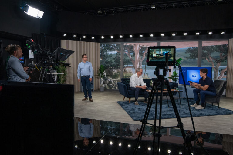 A behind-the-scenes view of a TV studio, showing a camera crew filming two men seated and talking on a set with a large window backdrop and a screen displaying a blue graphic. Another man stands nearby, observing.