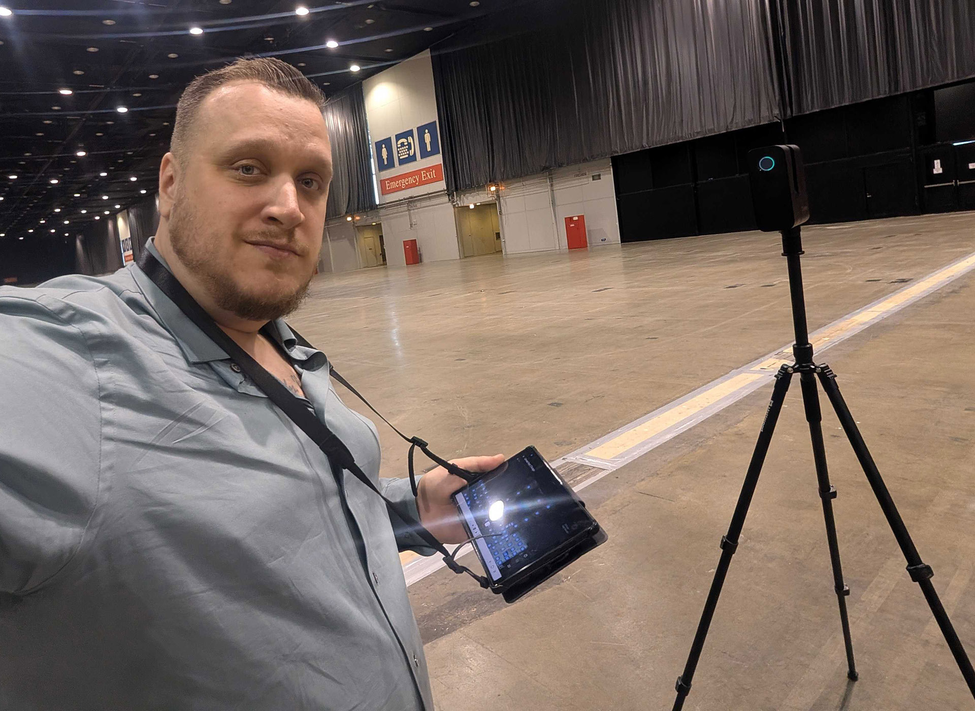 A man in a light blue shirt holds a tablet connected to a camera on a tripod in a large, empty convention center hall. Signs, emergency exit doors, and industrial lighting are visible in the background.