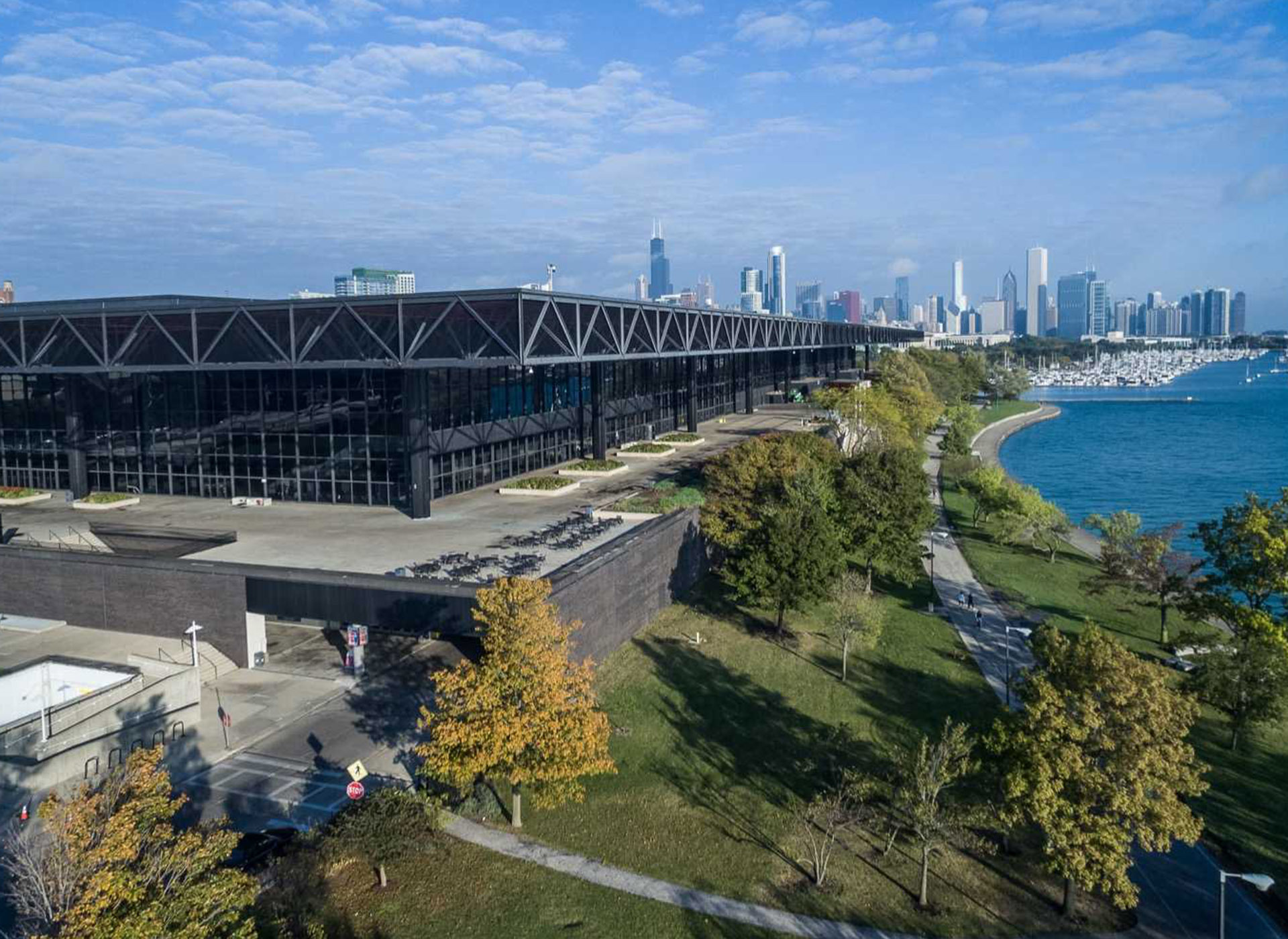 A large modern building with a black facade stands next to a tree-lined walkway along the edge of a lake, with a city skyline visible in the background under a partly cloudy sky.
