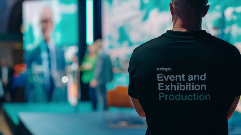 A person wearing a black Event and Exhibition Production shirt stands in the foreground, facing a brightly lit stage where people are gathered, with large screens displaying blurred images in the background.