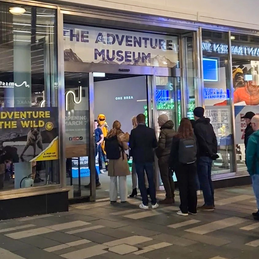 A group of people stands outside the entrance to The Adventure Museum, which has large glass windows and displays colorful posters promoting adventure exhibits.