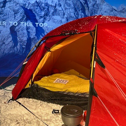 A red tent set up on snowy ground with its door open, revealing a yellow sleeping bag and a sign that says PLEASE DO NOT DISTURB inside. A cooking pot sits just outside, with snowy mountains in the background.
