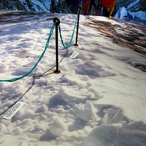 A snowy slope with a blue rope fence held by metal poles. Footprints are visible in the snow, and two small signs are placed on the ground. Two people with climbing gear appear in the background.