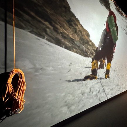 A coiled climbing rope hangs on a wall in front of a large photo showing a climber ascending a snowy, steep mountain slope with climbing gear and yellow boots.