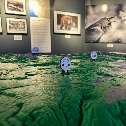 A close-up of a 3D topographic map with numbered audio guide markers. Framed wildlife photos and informational posters hang on the wall in the background of an exhibit space.