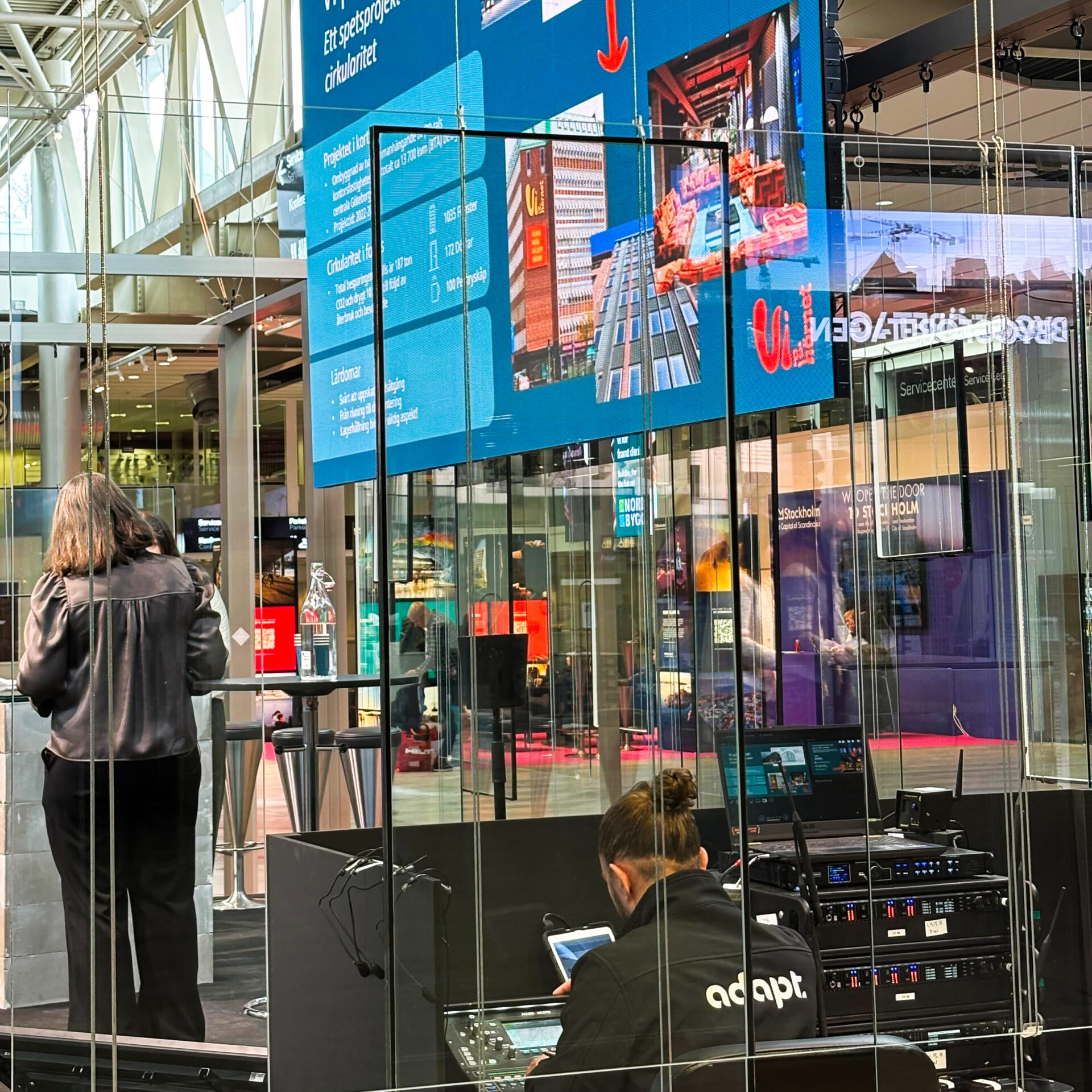 A woman stands on a stage near a podium, while a person labeled adapt audiovisual equipment. Large digital screens display graphics and text in a bustling indoor public space with glass walls.