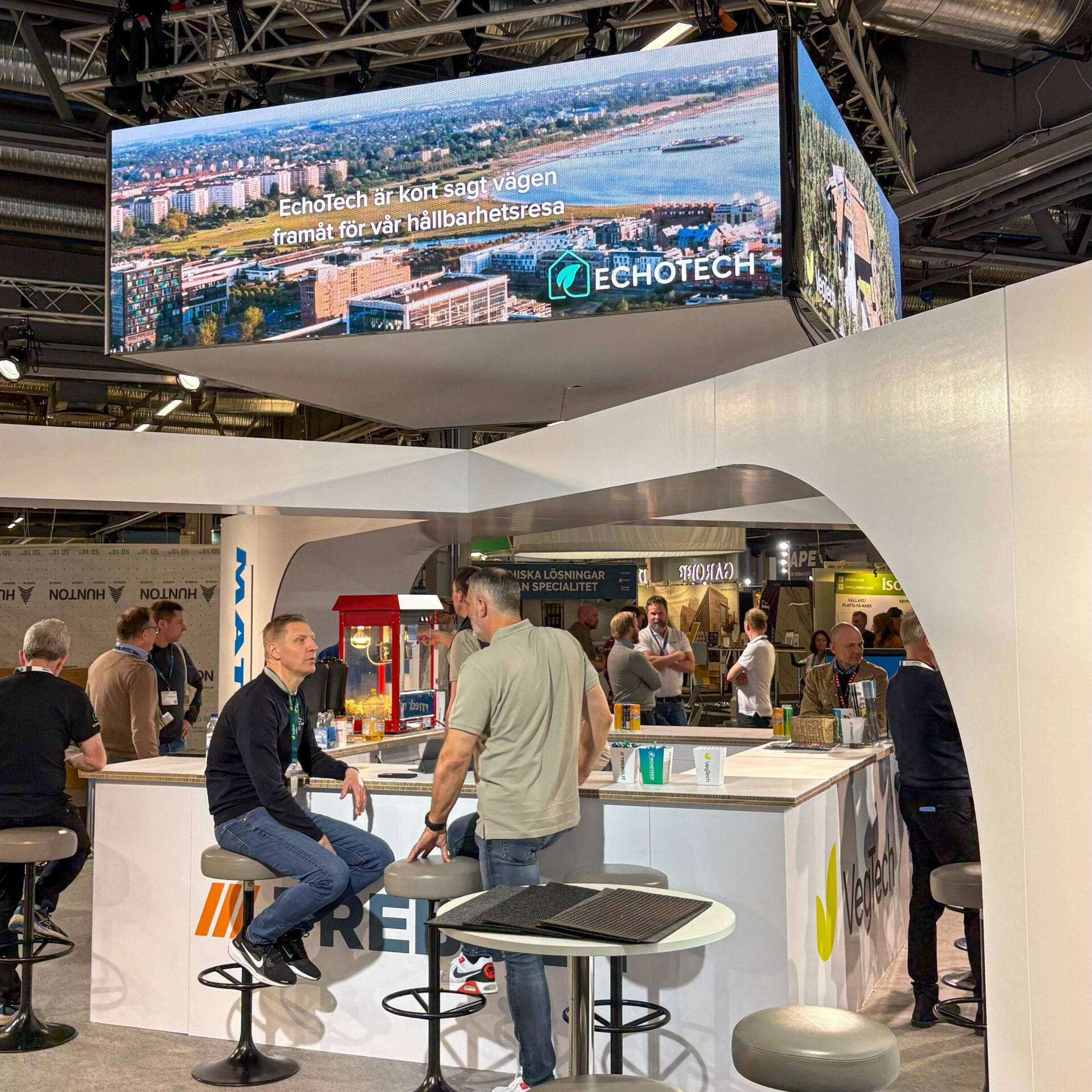 A trade show booth with people chatting around a counter. Above, a large screen displays a cityscape and the text “In short, ECHOTECH is the way forward on our sustainability journey,” along with the Echotech logo.