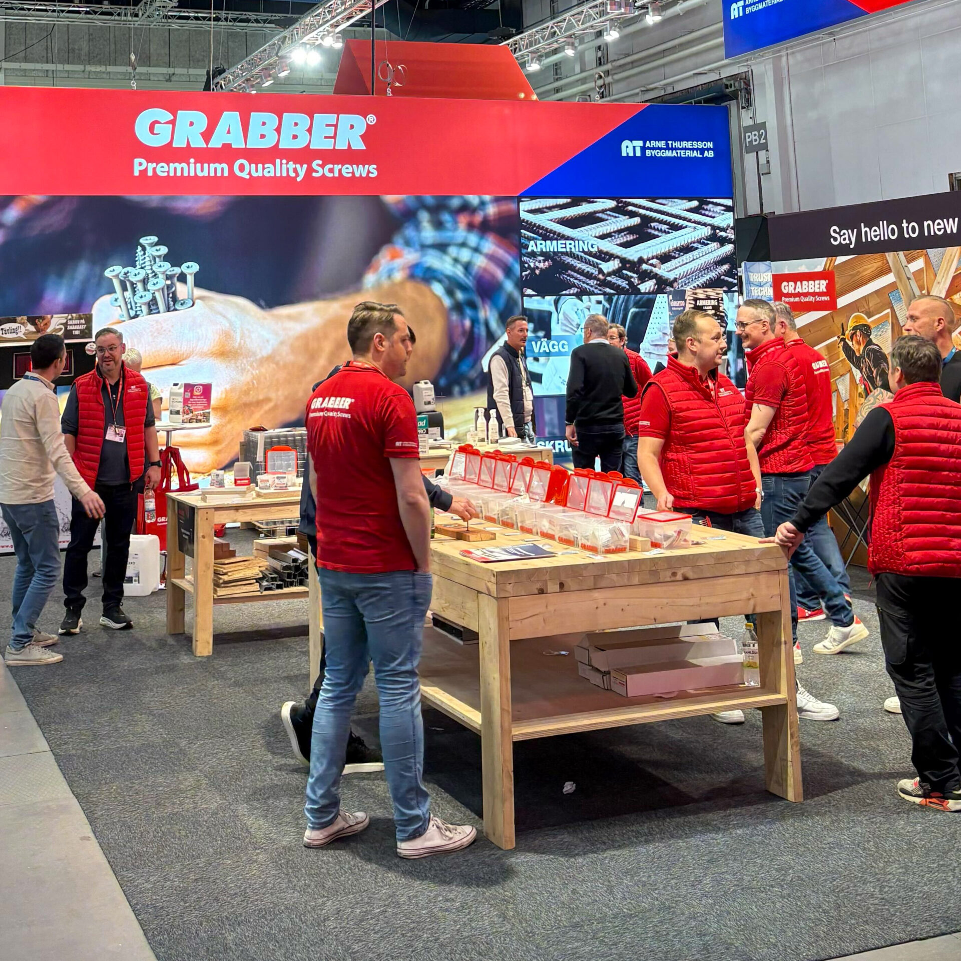 Several people are interacting at the GRABBER trade show booth, a company that specializes in premium screws. The booth features wooden tables displaying products and promotional materials, and staff in red vests are assisting visitors.