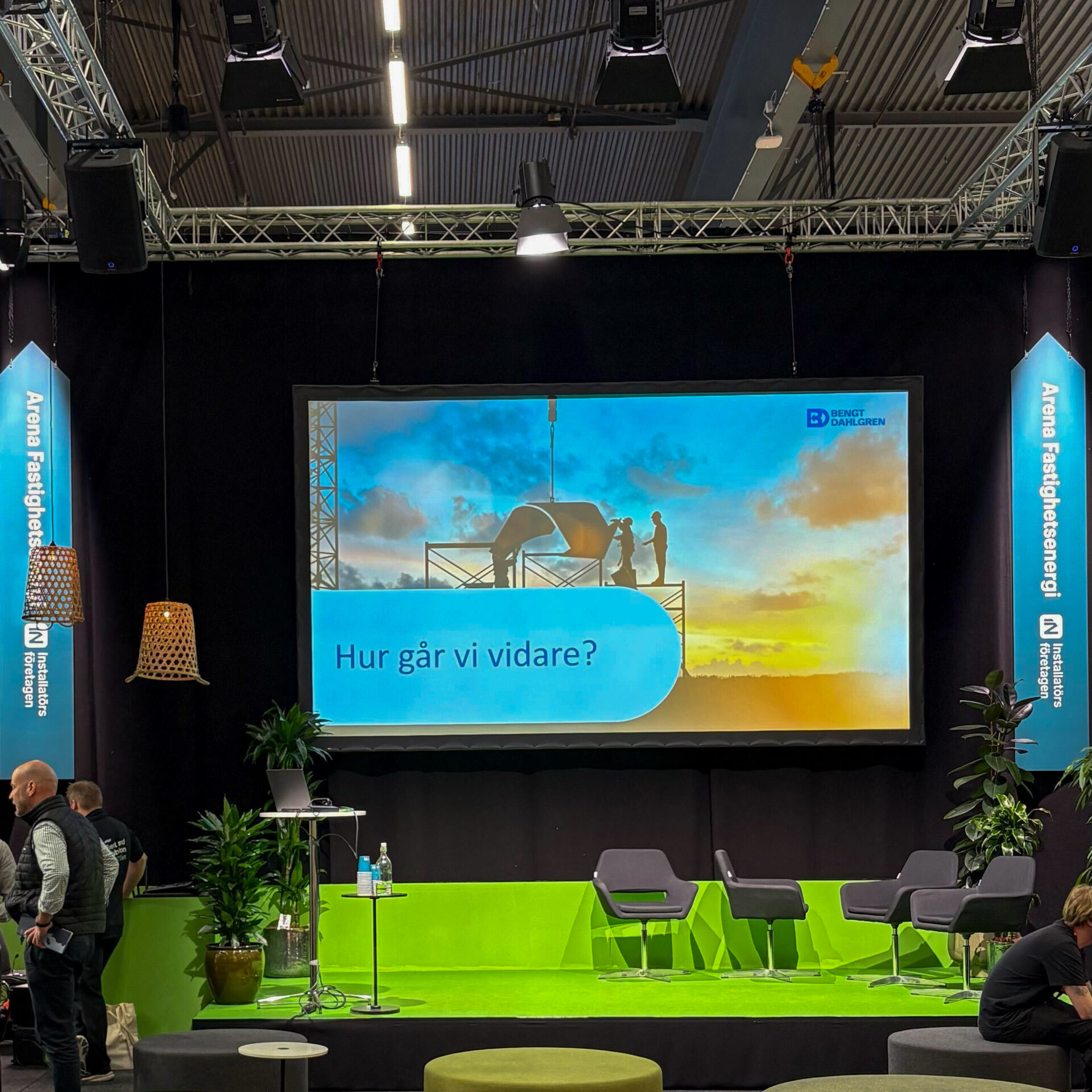 A stage with green flooring and chairs has been set up for a presentation. A large screen displays an image of two people on scaffolding at sunset, with the Swedish text "Hur går vi vidare?" (meaning "How do we move forward?").