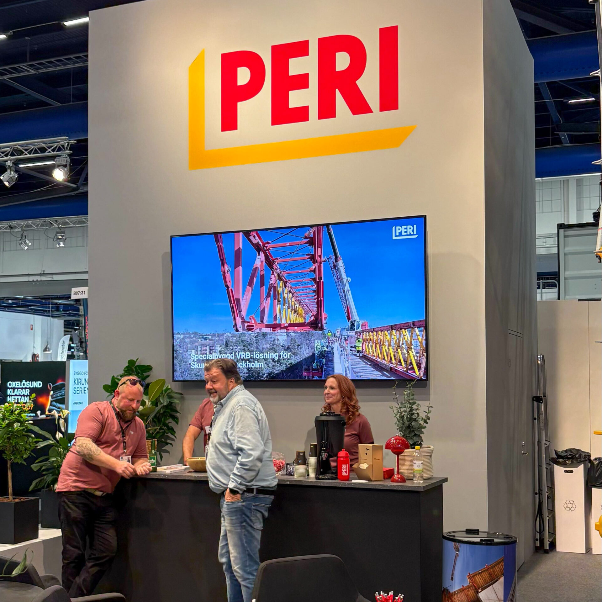 Three people are standing and talking at a booth with a large PERI logo above them. A screen displays an image of construction scaffolding. The booth features plants, coffee supplies, and a modern design.