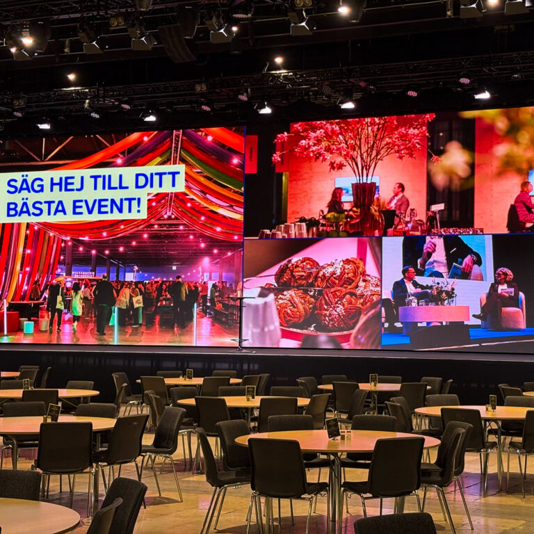 A large event hall with empty round tables and chairs, facing a bright screen displaying colorful event photos and the Swedish text “SÄG HEJ TILL DITT BÄSTA EVENT!”, which means “Say hello to your best event!”.