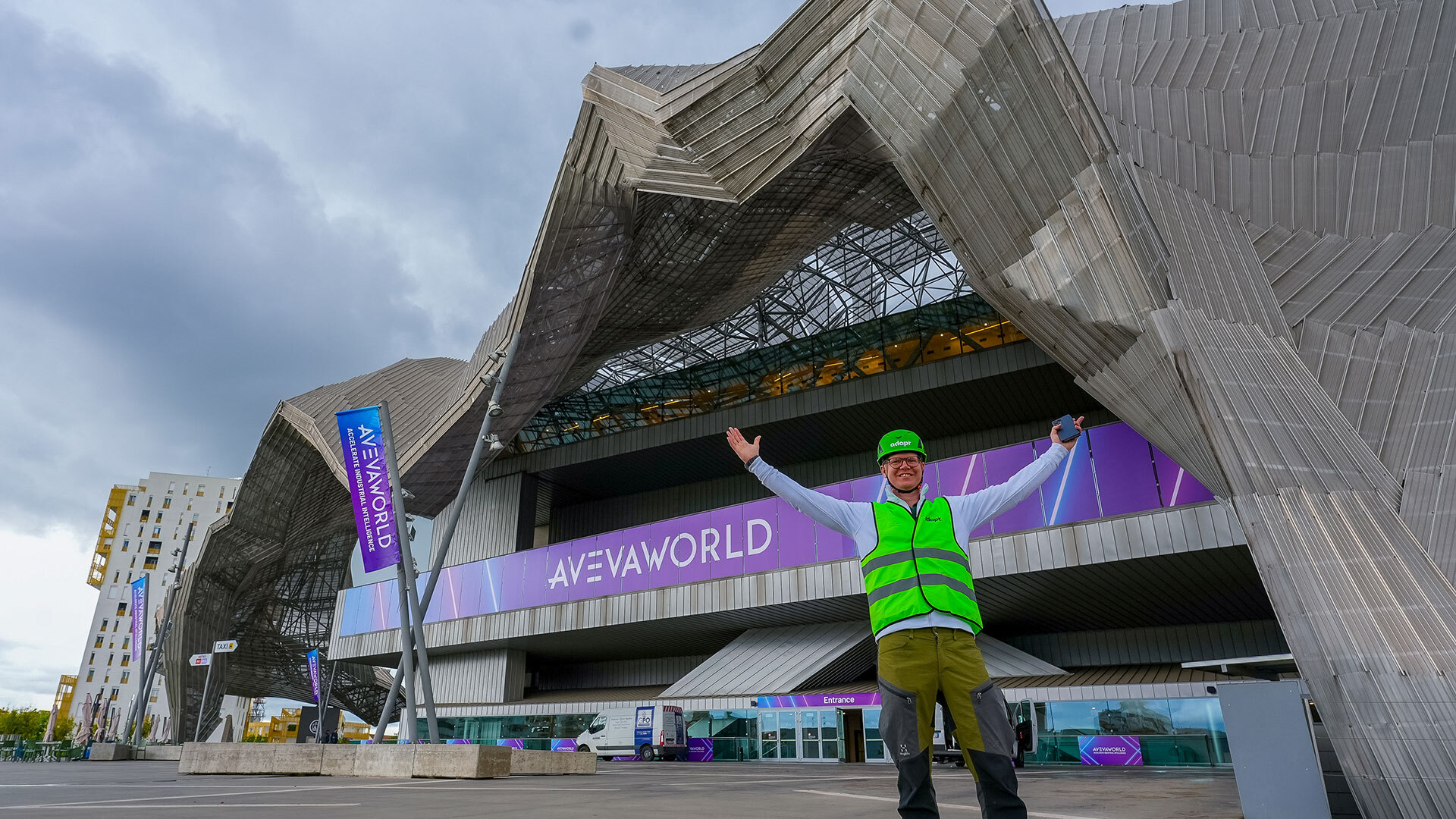 A person wearing a bright green safety vest and hard hat stands with arms raised in front of a modern building with angular architecture and signage that reads AVEVA WORLD.