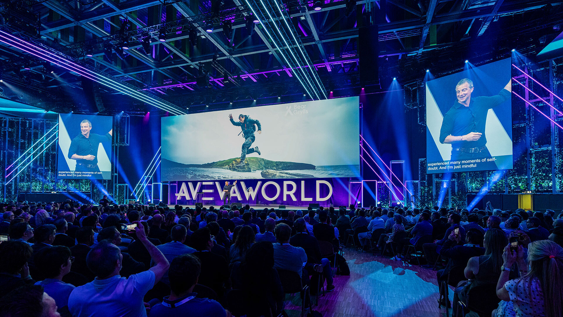 A speaker presents on stage at a large conference, with a screen showing an adventurer and the words AVEVA WORLD. The audience listens while two smaller screens display the speaker and a sign language interpreter.