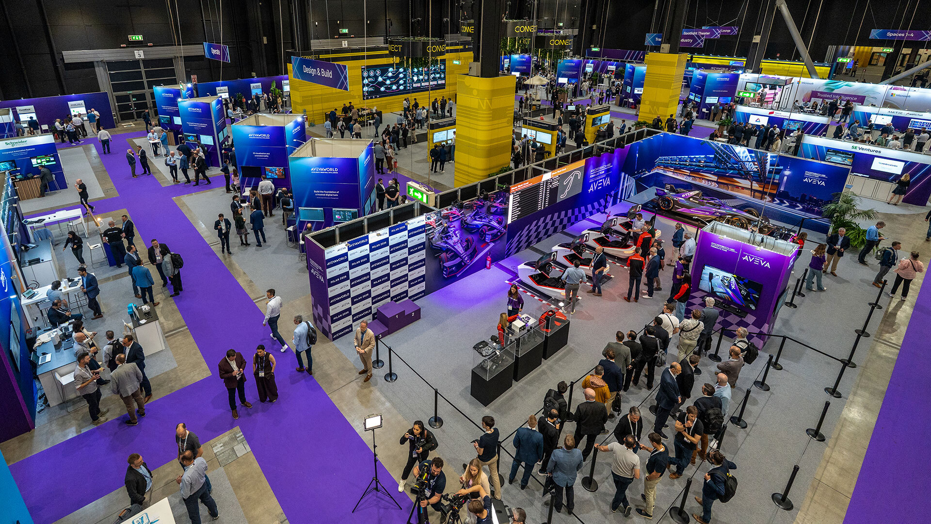 A large tech expo with numerous booths, display screens, and people walking, networking, and viewing exhibits, including a racing car display area. The venue features bright lighting and purple carpeting.