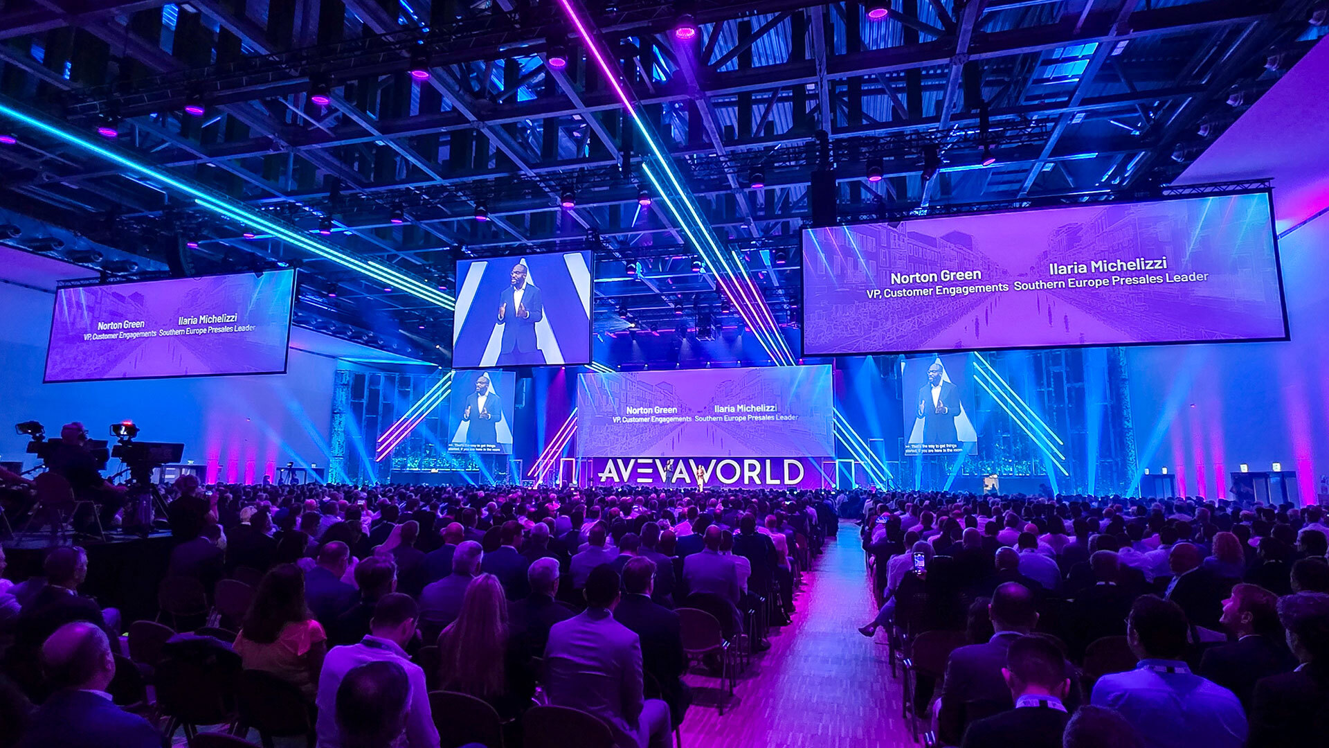 A large audience sits in a conference hall with vibrant blue and purple lighting, facing a stage where a speaker presents. Multiple large screens display the speaker and event information. The event is titled AVEVA World.