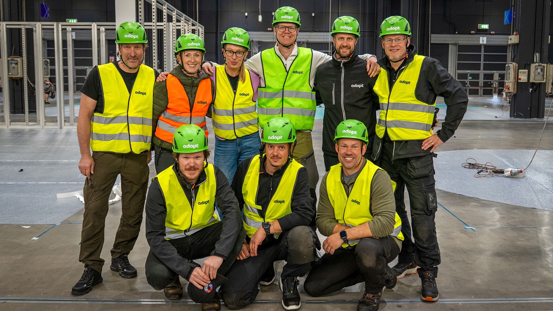 A group of ten people wearing green helmets and yellow or orange safety vests pose and smile together inside an industrial building under construction. Metal frames and equipment are visible in the background.