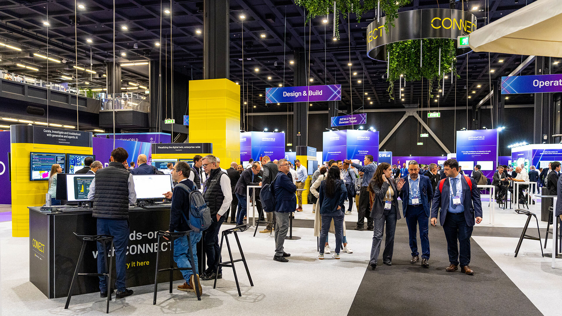 A busy convention hall with people interacting, large display screens, and colorful signs for sections like “Design & Build” and “Operate.” The space features modern decor, tall ceilings, and bright lighting.