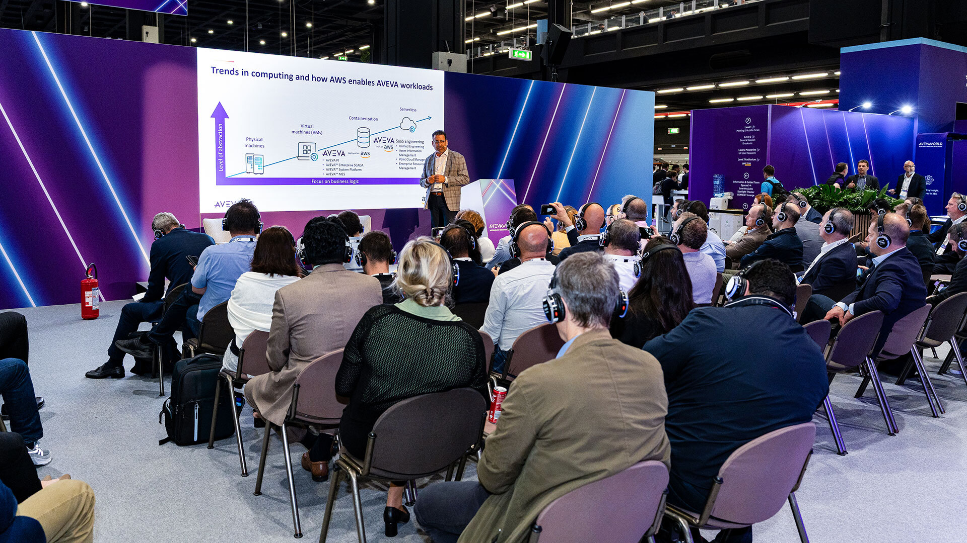 A speaker presents a tech-related slideshow to an audience wearing headphones at a conference. The presentation screen shows a diagram on trends in computing and ARM architecture. The room has modern decor with blue and purple lights.